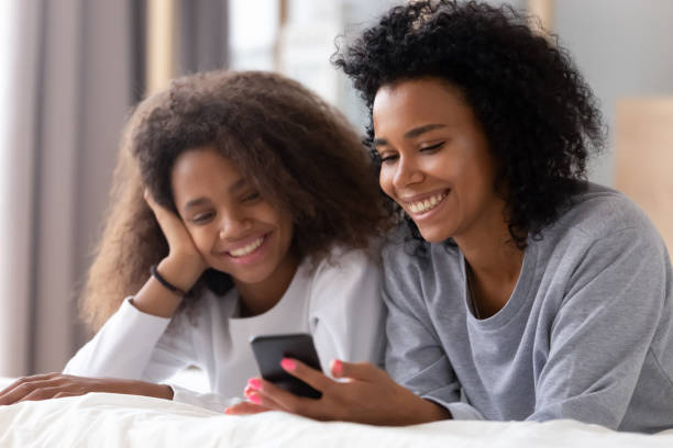 Happy african american mother and teen daughter using smartphone apps for shopping social media lying on bed, smiling black mom with teenage girl having fun watch videos on phone laughing in bedroom