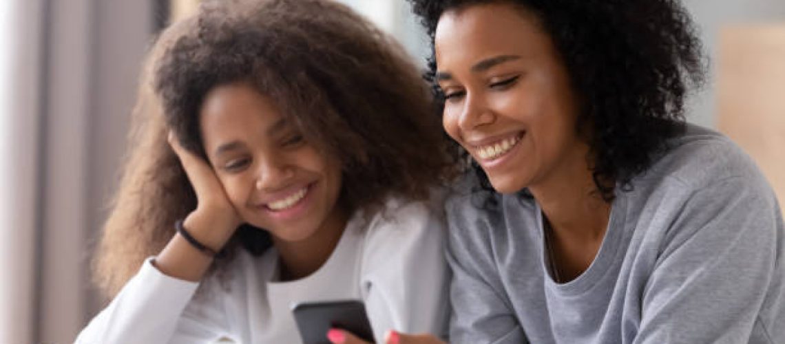 Happy african american mother and teen daughter using smartphone apps for shopping social media lying on bed, smiling black mom with teenage girl having fun watch videos on phone laughing in bedroom